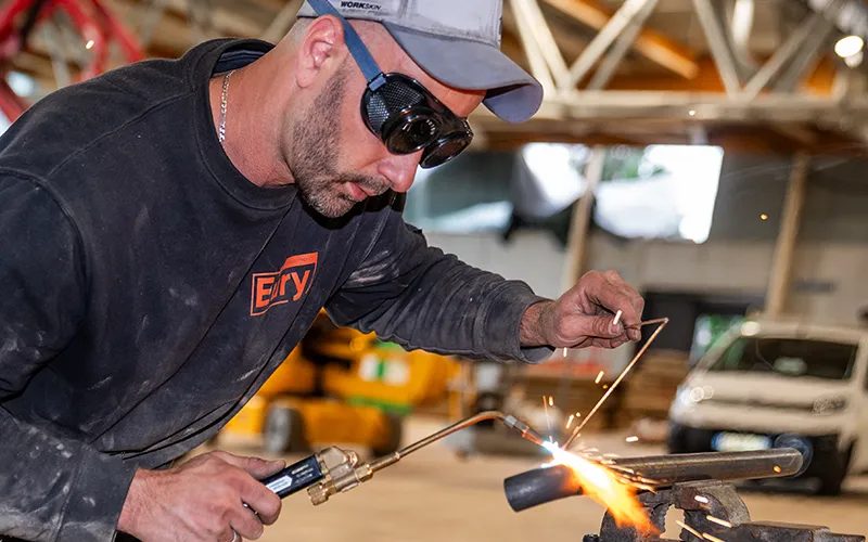 Technicien en t-shirt Eury, lunettes de protection, soudant un tube au chalumeau