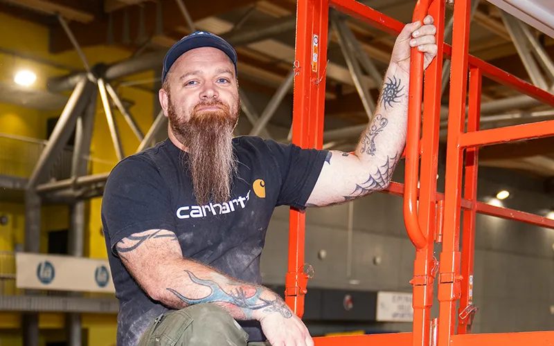 Technicien Eury barbu et tatoué, assis sur une nacelle orange